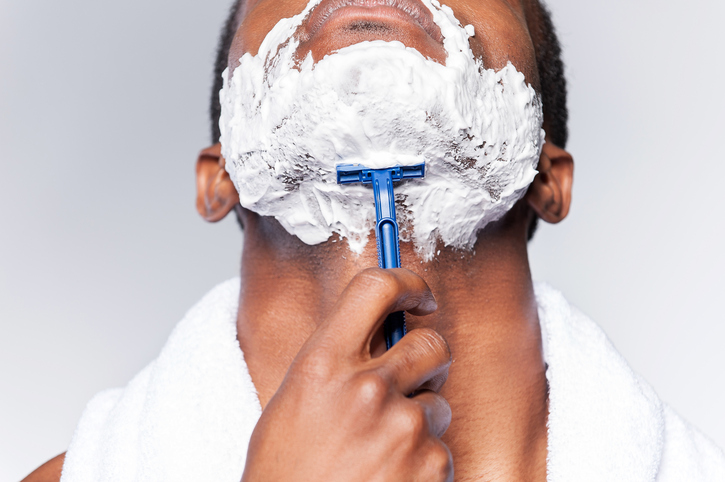 Close up of young African man shaving his face while standing against grey background