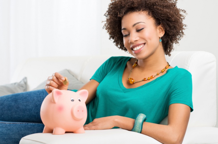 African American Woman on couch Putting Coin In Piggybank