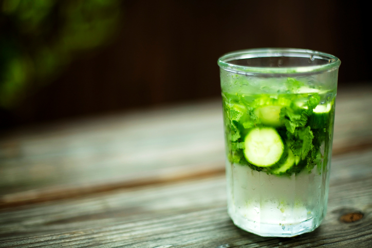 glass of iced water containing mint leaves and sliced cucumber placed on a rustic garden table