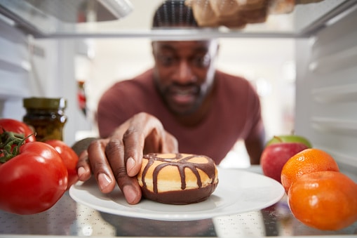 View Looking Out From Inside Of Refrigerator As Man Opens Door And Reaches For Unhealthy Donut