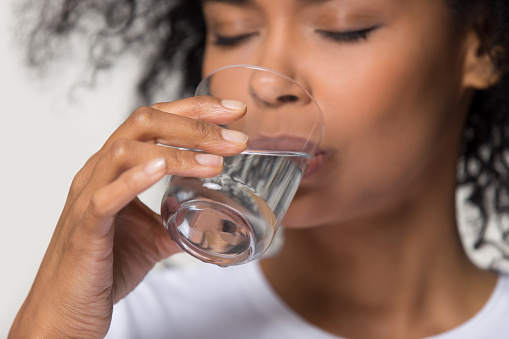 Close up focus on female hand holding glass african woman drinking still water having thirst healthy lifestyle body skin care hydration aqua balance regulation concept, studio shot on grey background